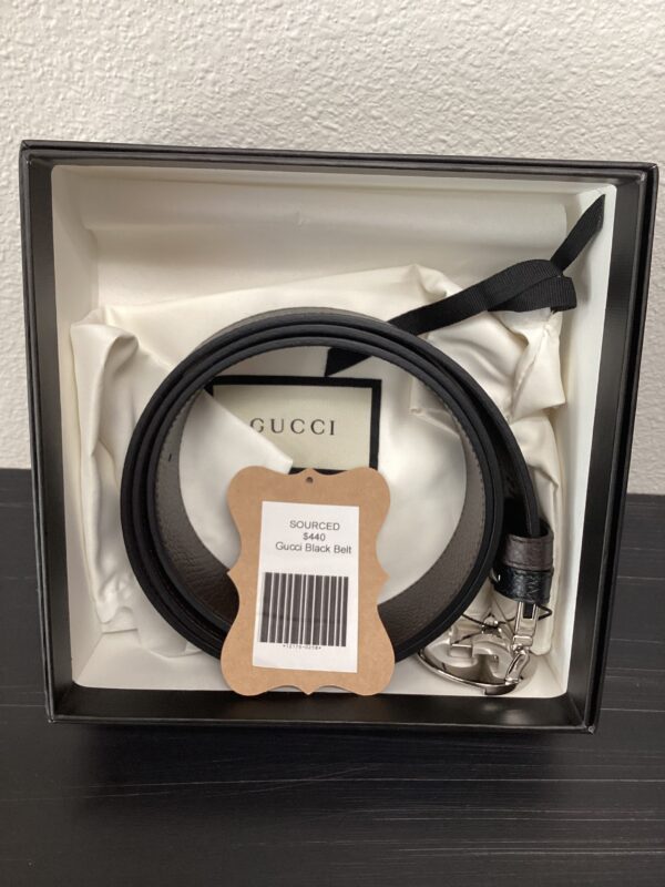 A gucci black belt with a silver buckle, displayed in a framed box with a label.