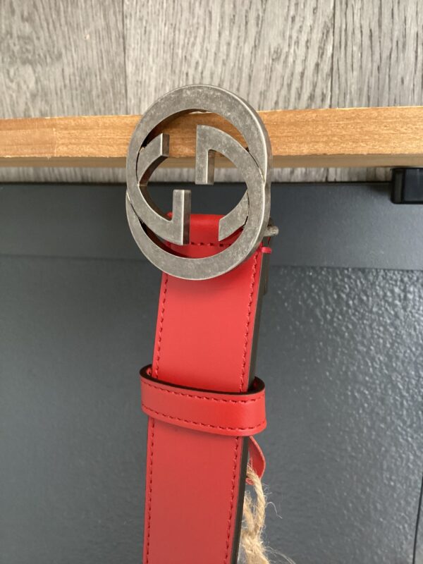 A Gucci red belt with a silver interlocking g buckle, hanging against a wooden panel and gray background.