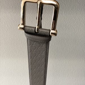 A close-up of a gucci belt with a gold buckle, hanging against a gray background. the belt features a patterned leather strap.