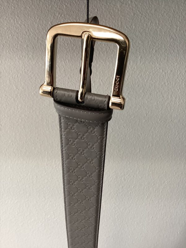 A close-up of a gucci belt with a gold buckle, hanging against a gray background. the belt features a patterned leather strap.