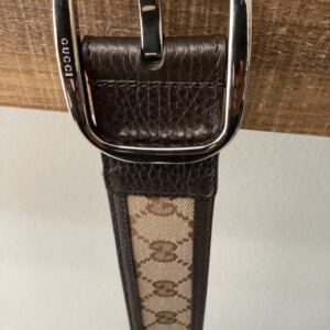 Close-up of a gucci belt with a silver buckle and logo on brown leather strap, partially visible against a wooden background.