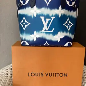 A LV MM Neverfull Bag with a blue and white pattern sits atop an orange branded box against a gray background.
