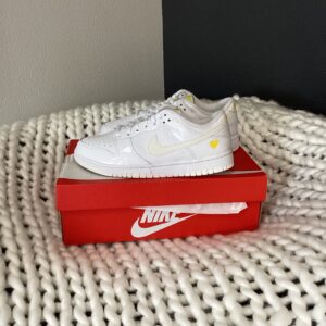 A white W Nike Dunk Low sneaker with a yellow heart emblem on a red W Nike Dunk Low shoe box, placed on a chunky, white knitted blanket against a black background.