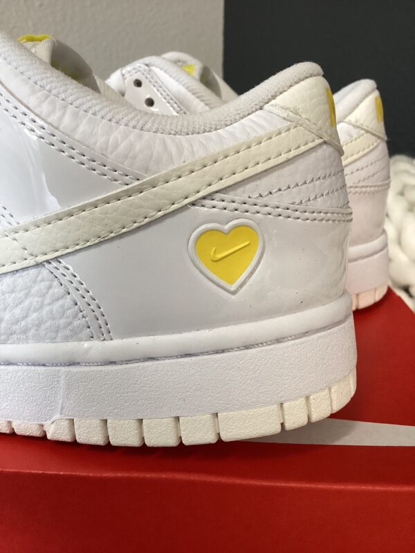 Close-up view of W Nike Dunk Low sneakers with a yellow heart design on the side and textured soles, displayed on a red surface against a grey background.