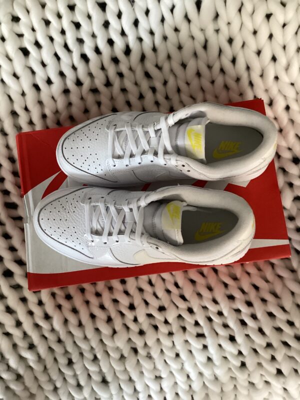 A pair of white W Nike Dunk Low sneakers with yellow accents displayed on a red shoebox, set on a textured white rug.