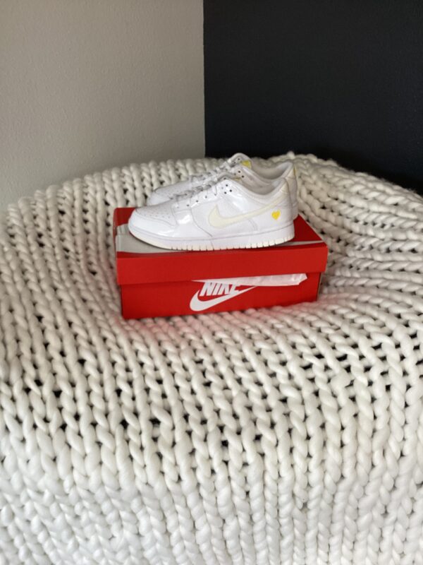 A white W Nike Dunk Low sneaker with a yellow accent rests on its shoebox on a textured white woven rug against a grey wall.