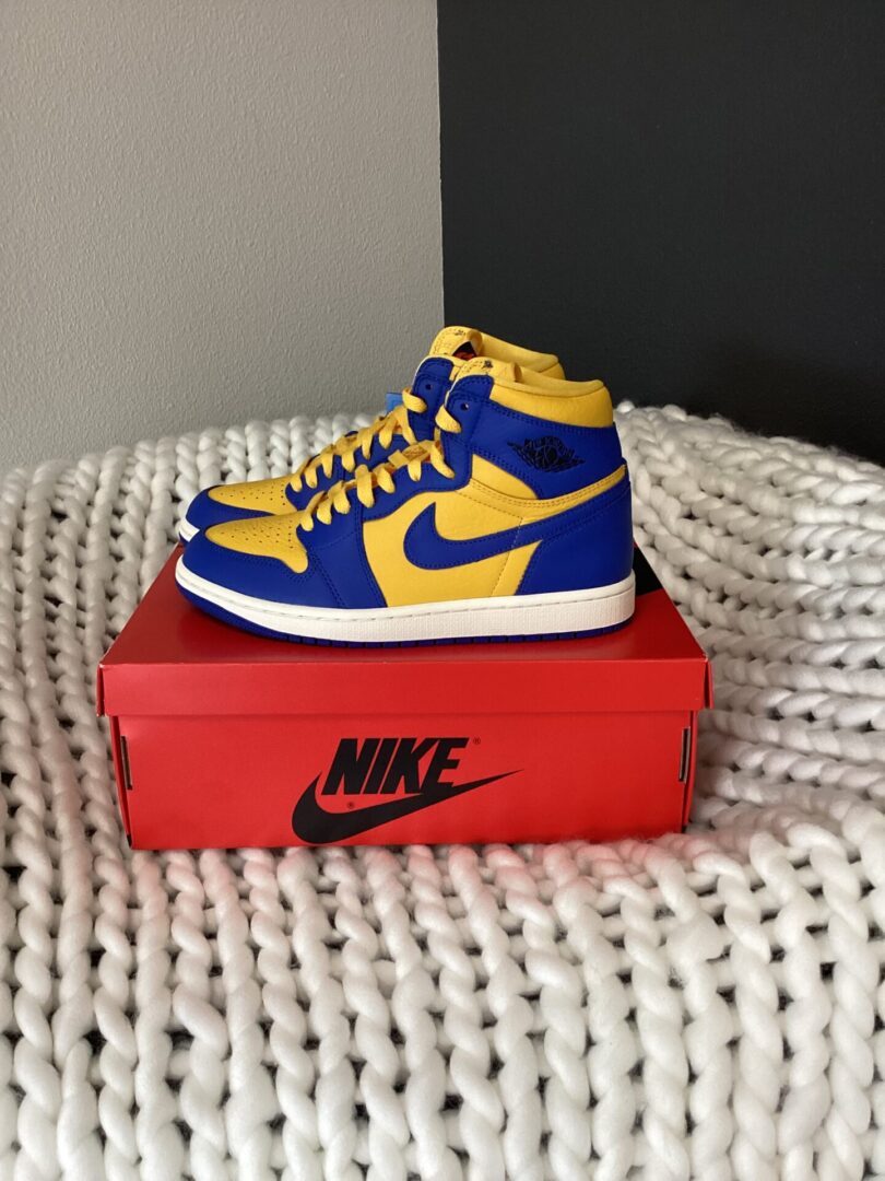 A pair of yellow and blue W Air Jordan 1 Retro sneakers sits on top of a red nike shoebox, placed on a white knitted blanket against a gray wall.