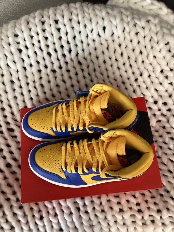 A pair of yellow and blue sneakers with red W Air Jordan 1 Retro logos, placed on a red shoebox over a white woven blanket.