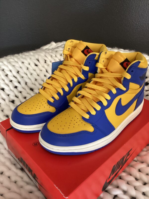 A pair of blue and yellow high-top W Air Jordan 1 Retro sneakers displayed on a red nike shoebox, placed on a white knitted surface.