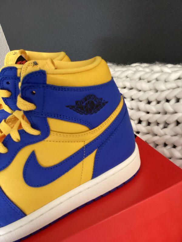 Close-up of a yellow and blue W Air Jordan 1 Retro sneaker with a logo on the ankle collar, displayed on a red box beside a white woven fabric.