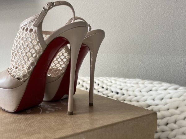 A pair of Christian Louboutin beige high-heeled shoes with red insoles on a wooden surface next to a white woven blanket.