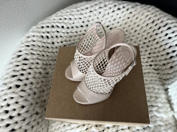 A pair of stylish Christian Louboutin beige high heeled sandals with a mesh design, displayed on a cardboard box against a white knitted background.