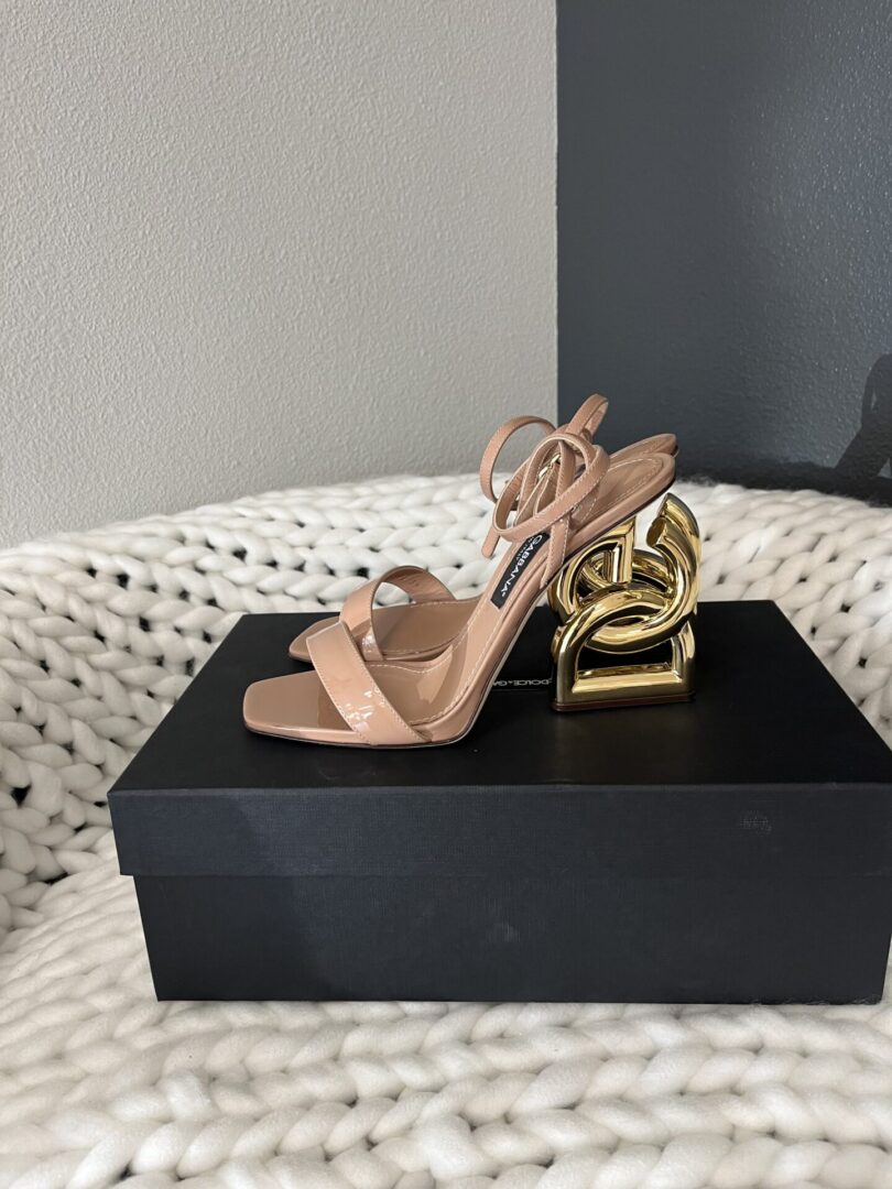 A pair of Dolce and Gabbana high-heeled sandals resting on a black shoebox beside a golden chain decor, all set on a white textured blanket.