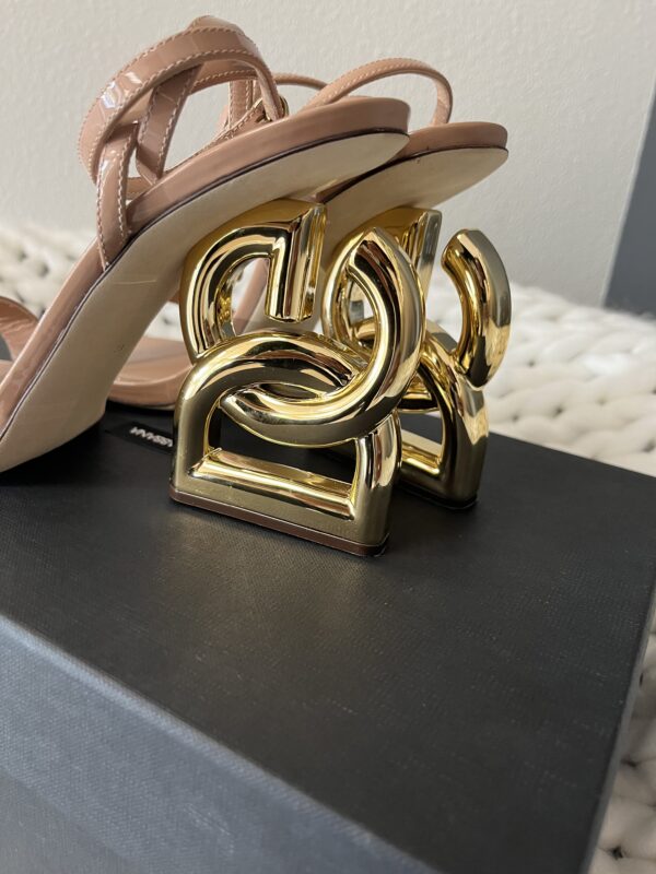 A pair of Dolce and Gabbana beige high-heeled sandals with a unique gold geometric heel design, displayed on a gray surface.