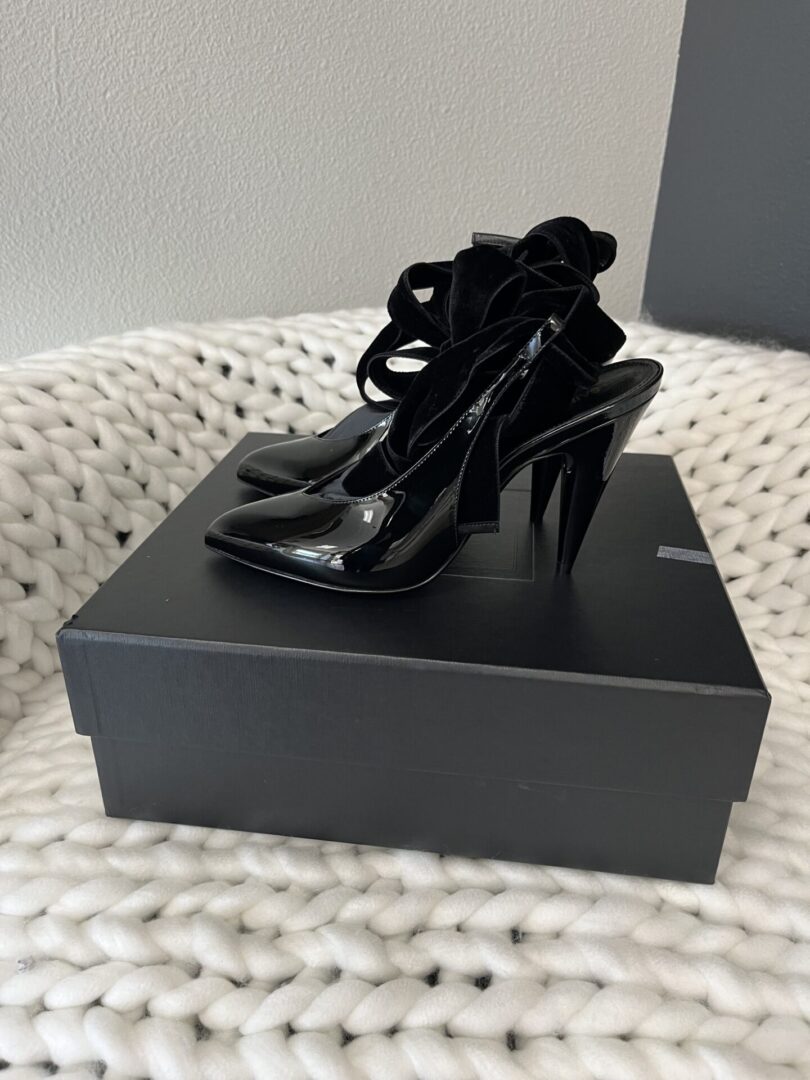 A pair of Saint Laurent Pumps with ribbon ties on top of a shoebox, placed on a white woven rug.