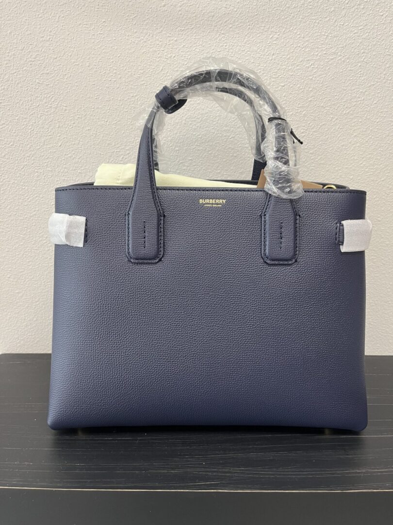 A new Burberry Top Handle Bag, in navy blue leather with protective plastic and foam on the handles and accents, displayed on a table against a white background.