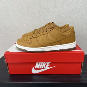A pair of brown Nike Dunk Low Wheat/Sail Black sneakers placed on top of their red and white Nike Dunk Low Wheat/Sail Black shoebox against a gray background.