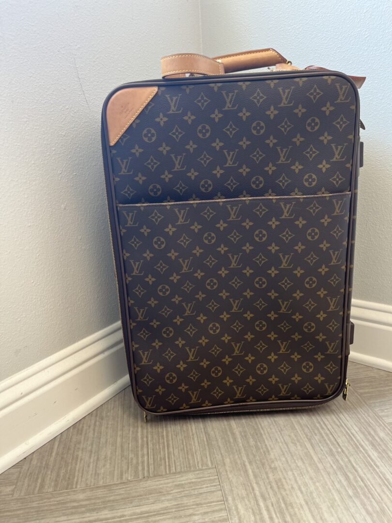A LV Rolling Luggage with a monogram design stands against a white wall on a gray tiled floor.