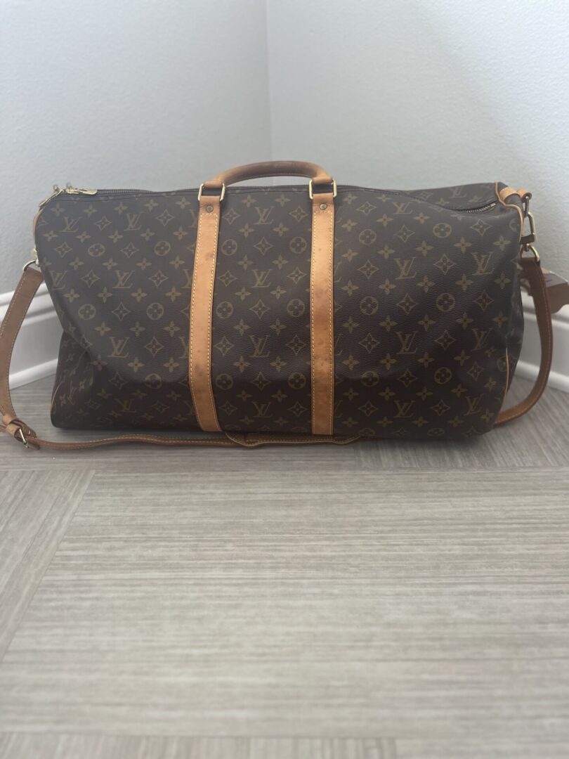 A Pre loved LV Dufflebag with the brand's iconic monogram pattern, featuring tan leather straps, on a gray floor against a white wall.