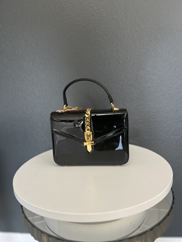 A small, Gucci black handbag with a gold clasp and handle, displayed on a white pedestal against a grey background.