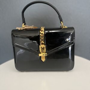 A glossy Gucci Black Handbag with a golden chain and clasp, displayed against a neutral gray background.