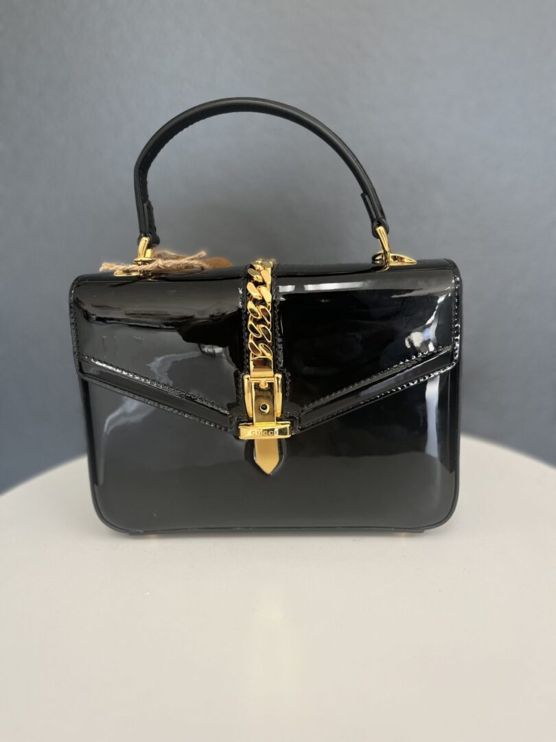 A glossy Gucci Black Handbag with a golden chain and clasp, displayed against a neutral gray background.