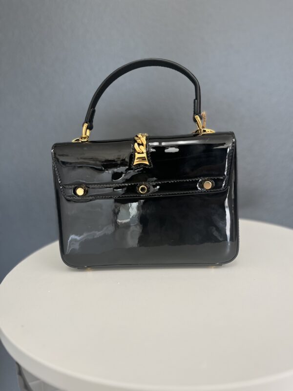 A shiny Gucci black patent leather handbag with gold hardware, standing on a white table against a gray background.