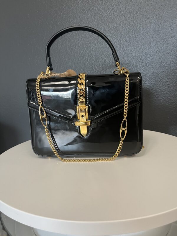 A Gucci Black Handbag with a gold chain strap and lock, placed on a white round table against a grey wall.