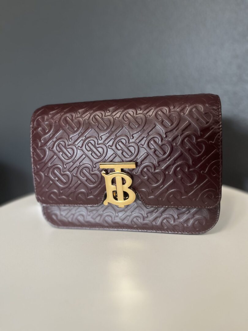 A dark red Gucci Black Handbag with an embossed pattern, featuring a prominent gold logo initials 'tb' on the front, set against a grey background.